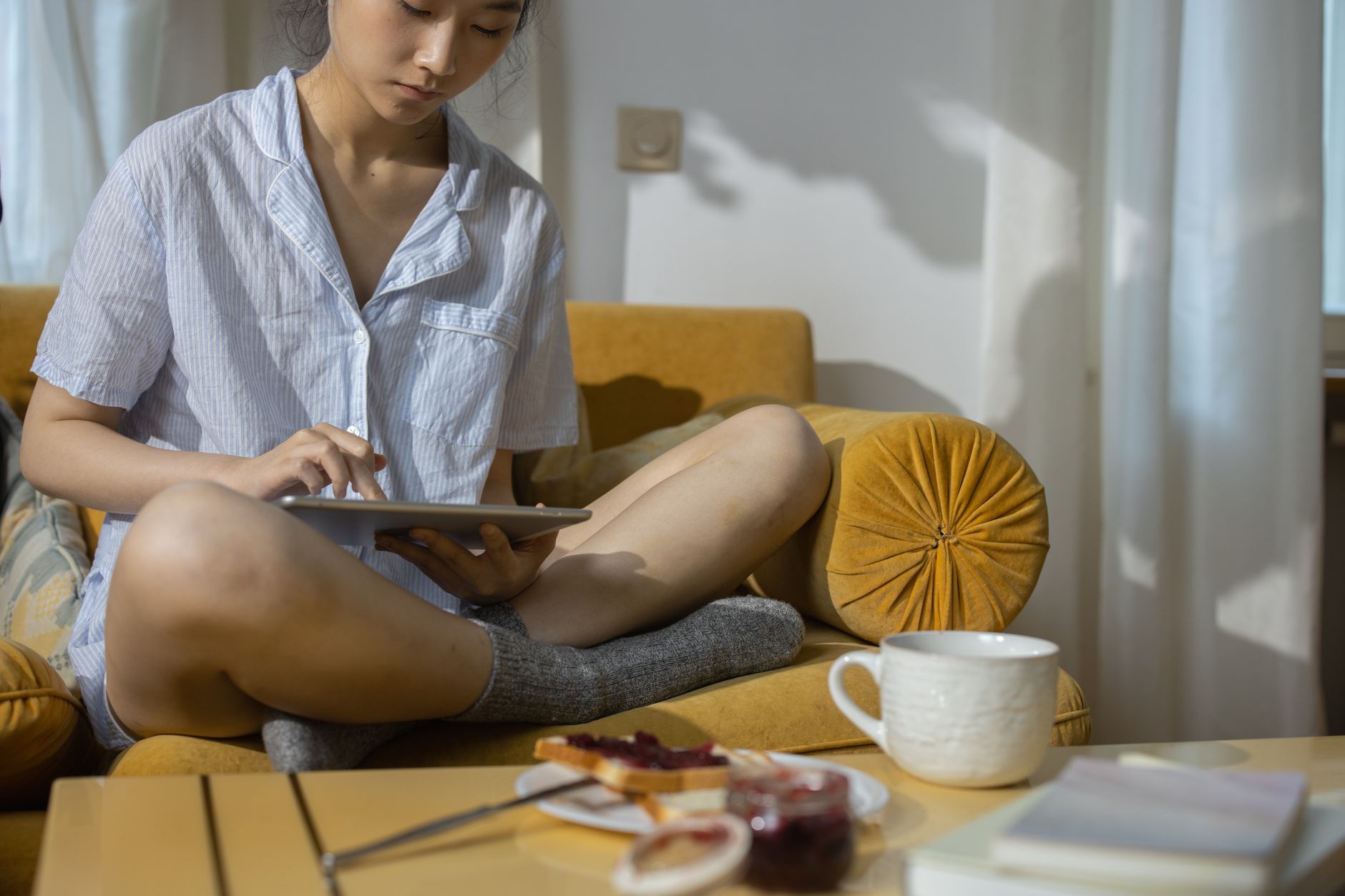 Une femme détendue en pyjama, assise sur un canapé avec des collations à proximité, qui utilise une tablette.